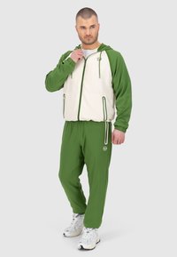 Giacca a vento verde e crema con tasche con zip e cappuccio, abbinata a pantaloni joggers verdi coordinati. Realizzata in tessuto liscio e leggero.