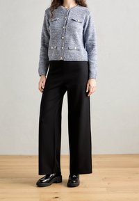 Pletený modrý kardigan se zlatými knoflíky a dvěma předními kapsami, v kombinaci s černými kalhotami širokého střihu a masivními černými botami na dřevěné podlaze.