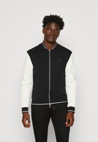 Chaqueta bomber en blanco y negro con cuerpo texturizado, mangas en contraste, puños acanalados y cierre de cremallera en el frente. Presenta un sutil logotipo en el pecho.