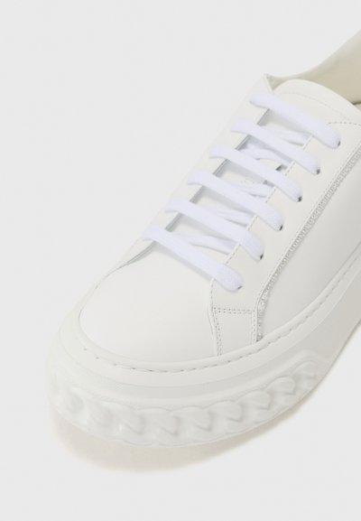 Vit lädersneaker med platta vita snören, rundad tå, texturerad vit sula med flätad design och accentdragkedja på sidan.