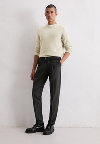 Hellbeige strukturierter Pullover, schwarze maßgeschneiderte Hose mit Falten und schwarze Schnürschuhe. Das Model steht vor einem neutralen Hintergrund.