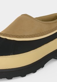 Gros plan d'une chaussure marron et noire à enfiler avec des coutures superposées et une semelle en caoutchouc noire texturée.