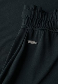 Tissu noir doté d'une taille élastique texturée et d'une étiquette en métal argenté. Matériau lisse et léger avec un léger éclat.