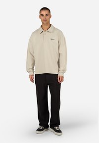 Bézs színű hosszú ujjú póló texturált anyagból, három gombos elülső résszel, fekete bő szárú nadrággal és fekete, fehér talpú sportcipővel párosítva.