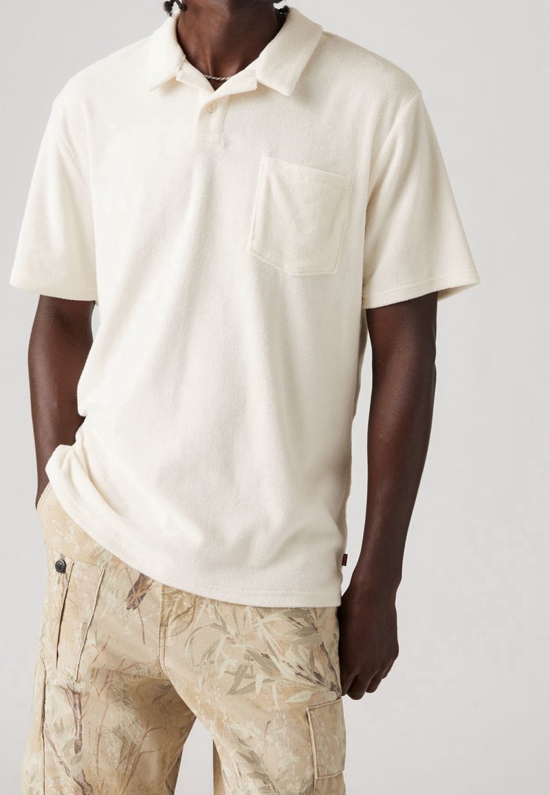Homme portant un polo crème à manches courtes avec une poche poitrine et un pantalon beige avec un motif camouflage feuillu.