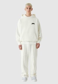 Biela mikina s čiernym logom, jemná tkanina, klokanie vrecko, spárovaná s bielymi teplákmi, ktoré majú štruktúrované detaily a manžety.