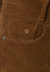 Bruin ribfluwelen stof met verticale ribbels, voorzien van een zak met een metalen knoop en stikdetails. Geborduurd logo vlakbij de zak.