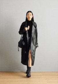 Svart lær trenchcoat med frontspalte, iført over en svart topp og teksturert skjørt. Kombinert med ankelstøvler og en liten håndveske.