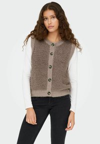 Braun strukturiertes Weste mit grünen Knöpfen und gerippten Kanten, über einem weißen Langarmhemd getragen; kombiniert mit schwarzen Denim-Hosen.