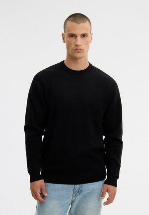 Un modelo masculino lleva un jersey negro de cuello redondo y unos vaqueros azul claro sobre un fondo blanco liso.