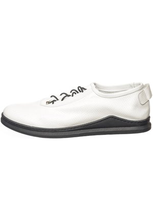 Weißer flacher Lederschuh mit schwarzer Sohle, schwarzen Schnürsenkeln, perforierter Struktur und einem Klettverschluss an der Ferse.