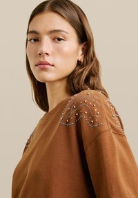 Sudadera marrón con cuello redondo, decorada con tachuelas plateadas en un patrón de olas en los hombros. Material de algodón con un acabado texturizado.