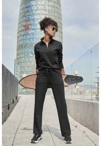Schwarze Zip-Up Sweatjacke mit rundem Logo, kombiniert mit passenden Sweatpants. Model hält ein Holzskateboard mit strukturiertem Oberflächen.