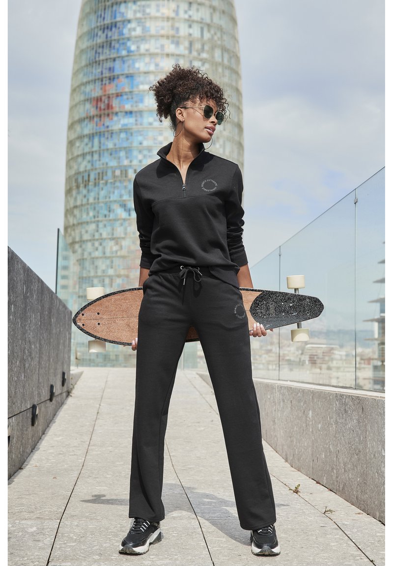 Schwarze Zip-Up Sweatjacke mit rundem Logo, kombiniert mit passenden Sweatpants. Model hält ein Holzskateboard mit strukturiertem Oberflächen.