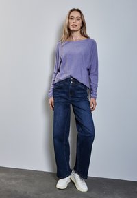 Camisetta a maniche lunghe viola abbinata a jeans a gamba larga blu scuro a vita alta e sneaker bianchi, in piedi contro un muro grigio chiaro.