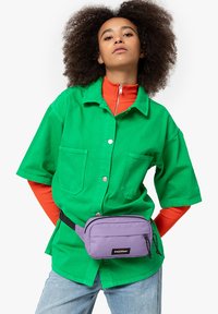 Vihreä lyhythihainen paita, jossa on kaksi taskua, oranssi korkea kaulus paita alla, ja violetti vyölaukku, jossa on mustia yksityiskohtia.