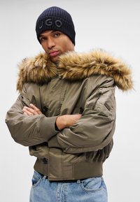 Hombre vestido con una chaqueta bomber verde con cuello de piel, gorro negro con el texto "HUGO" y jeans azules, con los brazos cruzados, mirando hacia adelante.