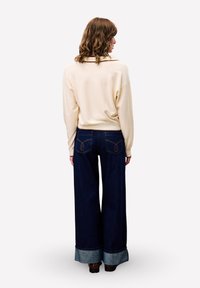 Vrouw met schouderlang krullend haar draagt een crème trui en wijdvallende donkerblauwe jeans met omslagen, staand met de rug naar de camera tegen een witte achtergrond.