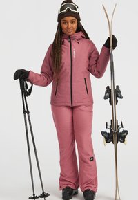 Roze skijack en broek, beide met een gestructureerd uiterlijk. Zwarte handschoenen en telescopische skistokken. Skies met zwarte bindingen, vastgehouden in één hand.