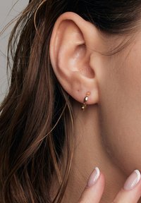 Złoty kolczyk w kształcie obręczy z skręconym wzorem i gładką teksturą, noszony na małżowinie usznej. Włosy częściowo zakrywają ucho.