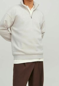 Sudadera de forro polar gris claro con un cuello alto, cierre de media cremallera y texto bordado "Original". Combinada con pantalones marrones.
