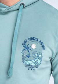 Világoskék kapucnis pulóver, amely pálmafák és hullámok grafikájával díszített. Fém végű húzózsinórral és "Surf Riders Hawaii" felirattal rendelkezik.
