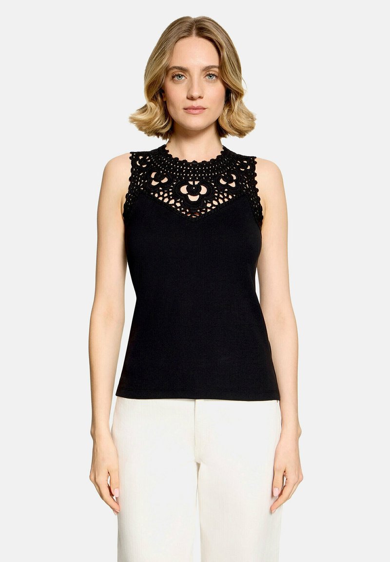 Femme aux cheveux blonds courts portant un haut noir sans manches avec des détails en dentelle et un pantalon blanc, debout devant un fond uni.