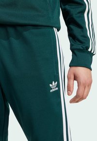 Grüne Sport-Trainingshosen mit weißem Drei-Streifen-Design. Verfügt über einen weichen Stoff, einen elastischen Bund und ein kleines weißes Adidas-Logo.
