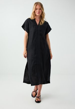 Blonde Frau, die ein lockeres schwarzes knielanges Kleid mit kurzen Ärmeln und schwarzen Sandalen trägt, steht vor einem einfarbigen hellen Hintergrund.