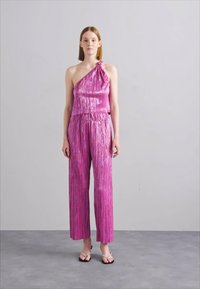 Combinaison brillante rose à une épaule, avec une texture plissée et des jambes larges. Portée avec des tongs noires simples.