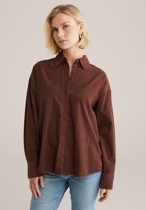 Femme blonde portant des boucles d'oreilles en or, une chemise marron à boutons avec des manches longues et un jean bleu clair, se tenant devant un fond uni.