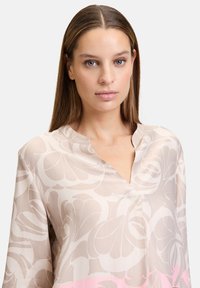 Leichtes, gemustertes Blusenshirt in Beige mit rosa Akzenten; zeichnet sich durch einen V-Ausschnitt und lockere Passform aus. Der Stoff hat eine glatte, weiche Textur.
