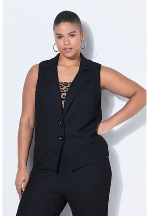 Itsevarma nainen, jolla on lyhyet hiukset, pukeutunut mustaan teksturoituun hihattomaan bleiseriin ja sopiviin housuihin leokuviollisen topin yli, poseeraa käsi lantiolla.