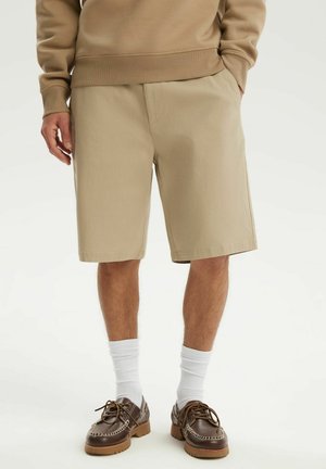 Mężczyzna ubrany w beżowe szorty, brązową bluzę, białe skarpetki oraz brązowe skórzane buty na sznurówki, stojący na białym tle.