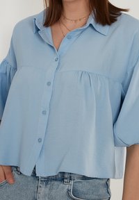 Blusa azul clara confeccionada en una tela suave, con un cierre de botones en la parte delantera, mangas sueltas y detalles fruncidos debajo del busto. Diseño ligeramente cropped.
