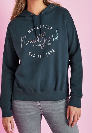 Žena v tmavě zelené mikině s nápisem „Manhattan New York Madison Avenue NYC Est. 1973“ v kombinaci se světle šedými džínami na růžovém pozadí.