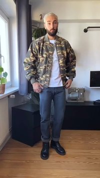 Homem com cabelo e barba descoloridos veste uma jaqueta camuflada, camisa branca, jeans e botas pretas, em pé numa sala com uma máquina de costura e plantas.
