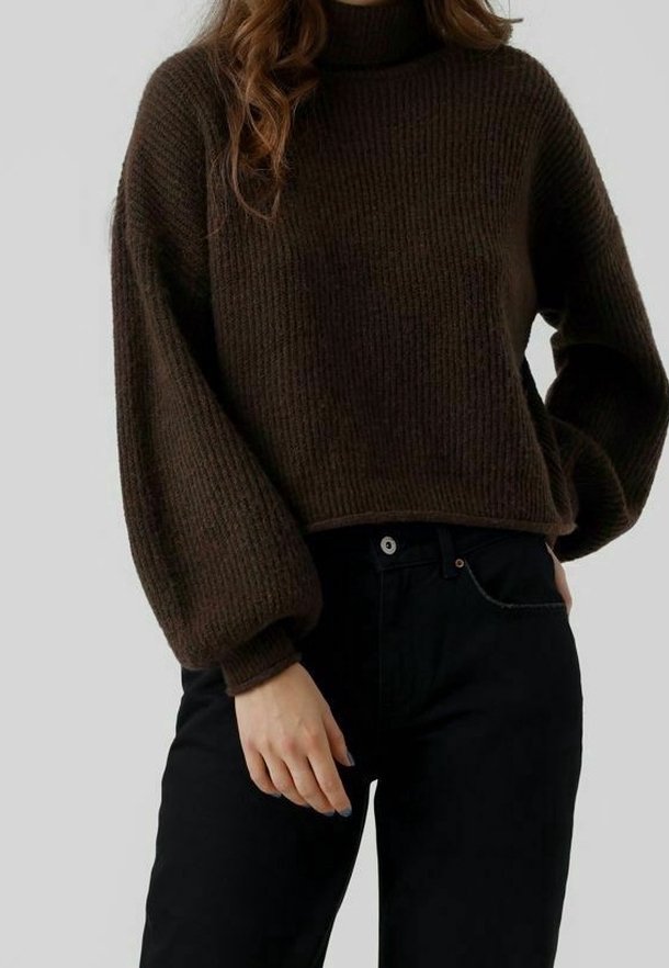 Femme portant un pull à col roulé côtelé marron foncé avec des manches bouffantes et un pantalon noir taille haute, debout avec une main dans la poche.