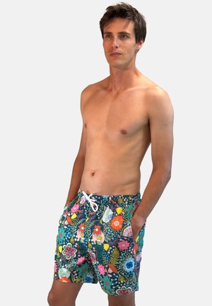 Homme torse nu debout, les mains dans les poches, portant un short de bain fleuri coloré avec un cordon, sur un fond uni clair.
