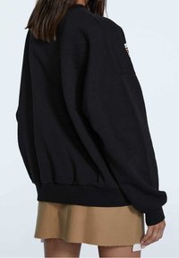 Sudadera negra de gran tamaño hecha de material suave, con puños y dobladillo acanalados, y un pequeño parche con el logo en el hombro izquierdo.