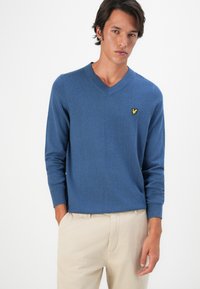 Uomo che indossa un maglione blu a V con un piccolo logo giallo sul petto, abbinato a pantaloni beige, in piedi di fronte a uno sfondo bianco semplice.