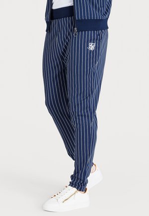 Donkerblauwe gestreepte joggerbroek met elastische tailleband, zijzakken en geborduurd logo, gecombineerd met witte sneakers met gouden accenten.