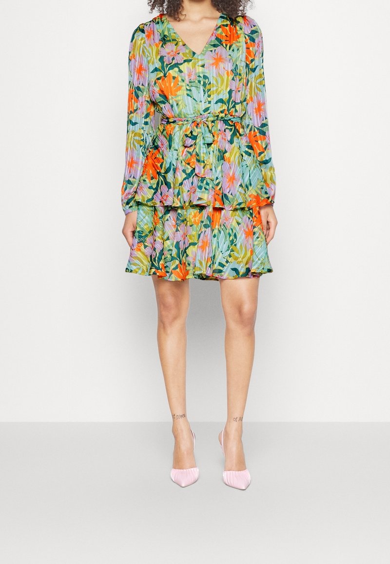Femme portant une robe fleurie plissée à manches longues avec un col en V et un ourlet à volants, associée à des chaussures à talons hauts rose clair.
