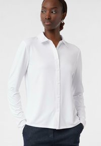Camicia bianca a maniche lunghe con colletto, realizzata in un tessuto morbido e leggermente elastico. Chiusura con bottoni sul davanti.