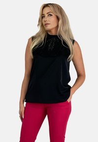 Czarny bezrękawnik z falbaniastym kołnierzykiem i ozdobnym dekoltem, połączony z dopasowanymi różowymi spodniami. Top ma gładką fakturę.