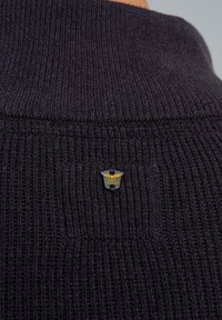 Pull en maille violet foncé avec un design en côtes et un patch logo bien visible au niveau du col. La texture semble douce et épaisse.