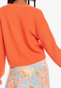 Helles orangefarbenes Strickpullover mit einer strukturierten Musterung und einem rundem Halsausschnitt, kombiniert mit einem floral bedruckten Rock in Blau, Orange und Weiß.