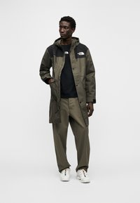 Αδιάβροχο μπουφάν σε ελιά πράσινο χρώμα με μαύρο επάνω τμήμα, μπροστινές τσέπες και κουκούλα με κορδόνι, συνδυασμένο με παντελόνι σε ίδιο ελιά πράσινο χρώμα και λευκά αθλητικά παπούτσια.