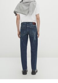 Tummansiniset farkut suoran lahkeisen leikkauksen kanssa, näkyvät takataskut ja ruskea nahkalaatta vyötäröllä. Siisti, minimalistinen muotoilu.