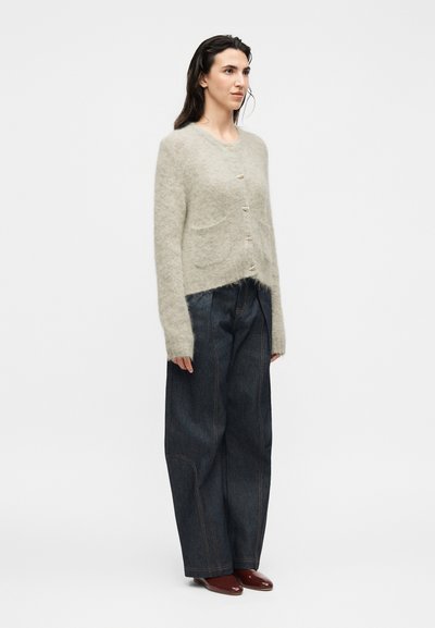 Femme debout portant un cardigan gris clair en matière duveteuse boutonné, un jean large en denim foncé et des chaussures pointues marron sur un fond blanc uni.